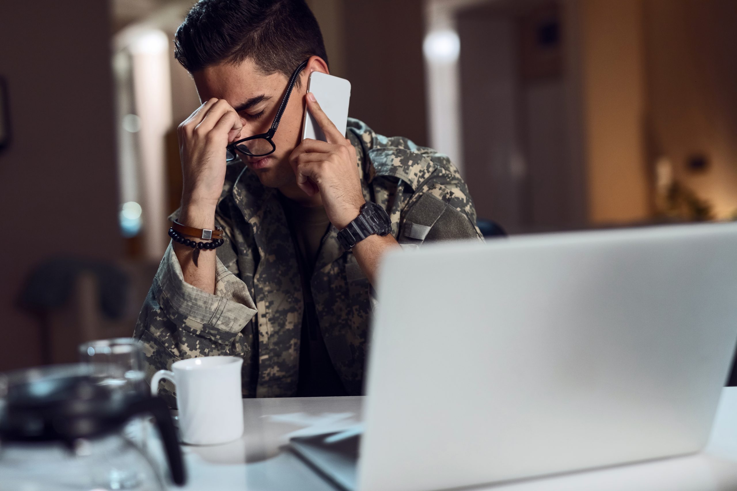 Young army soldier having headache while talking on mobile phone and working at desk office.