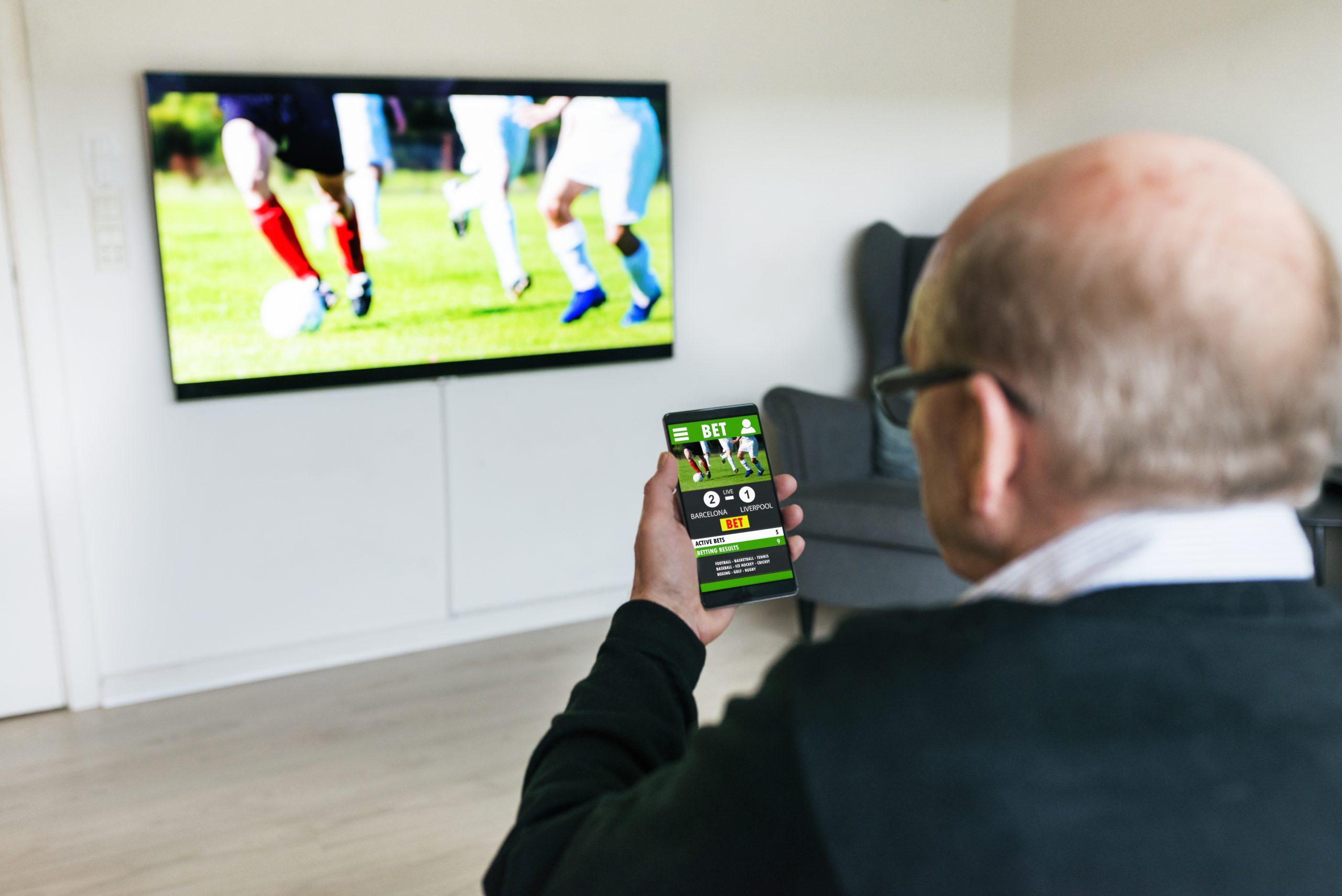 Senior man sits in front of flat screen television. He holds a mobile phone which displays a betting app and a current soccer match with live results. There is an option to bet on the match.